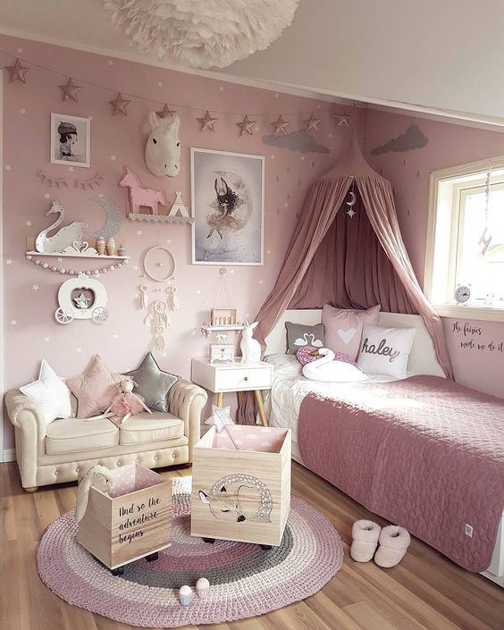 دکوراسیون اتاق خواب کودک دختری با دیوار صورتی که تور بالا تخت صورتی، مبل کوچک کرم، مجسمه اسب تک شاخ و دکوری های دیواری دارد